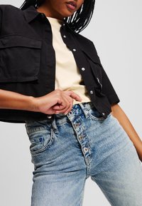 Camicetta nera tagliata con due tasche a toppa, abbinata a jeans a vita alta in lavaggio chiaro con una chiusura a cinque bottoni e lievi segni di usura.