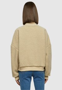 Beige Fleece-Jacke mit weich strukturierter Oberfläche, oversized Fit und gerippten Bündchen. Über blauen Jeans getragen, von hinten betrachtet.