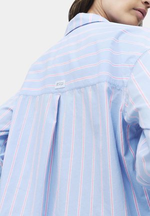 Frau trägt ein hellblaues Hemd mit rosa und weißen vertikalen Streifen, gezeigt von hinten und seitlich, mit einem kleinen Etikett am oberen Rücken.