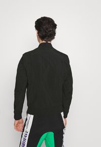 Černá bomber bunda s hladkou strukturou, s stojáčkovým límcem a žebrovanými manžetami, zobrazená zezadu na bílém pozadí.