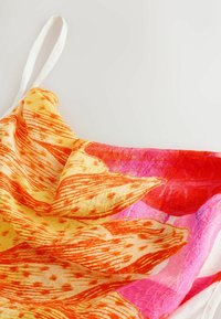 Robe à bretelles avec motifs de feuilles orange et jaune et accents roses sur un tissu léger légèrement froissé sur fond blanc.