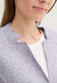 Lavendelfärgad jacka i en texturerad tyg med öppen krage och råkantade detaljer, lagd över en vit skjorta. Mjuk och lätt i utseende.