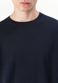 Pull en maille bleu marine, fabriqué dans un tissu doux, avec un col rond côtelé et un logo brodé discret sur la poitrine.