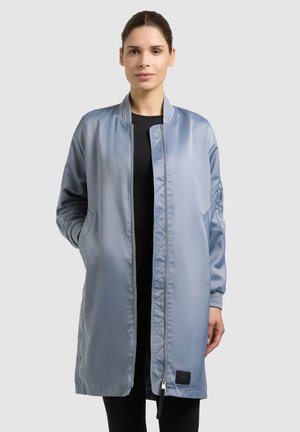 Frau steht da und trägt eine lange, hellblaue Satin-Bomberjacke über schwarzer Kleidung, die Hände locker neben dem Körper, vor einem einfarbigen hellgrauen Hintergrund.