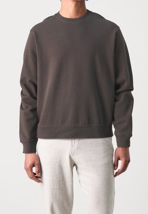 Person, die einen dunkelbraunen Langarm-Pullover mit Rundhalsausschnitt und hellbeige Hosen trägt, vor einem einfarbigen Hintergrund steht.