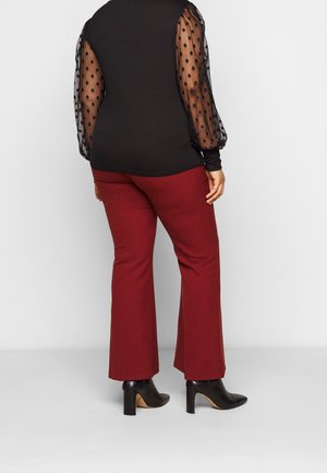 Kobieta w czarnej prześwitującej bluzce w groszki z długim rękawem, czerwonych dopasowanych spodniach i czarnych botkach na obcasie, stojąca na białym tle.