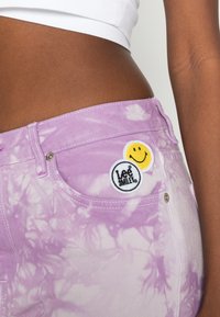 Jeans de ganga tie-dye roxos com duas remendos circulares: um sorriso amarelo e um logótipo "Lee Smile" em preto e branco.