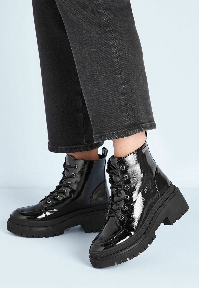 Bottes noires brillantes en cuir verni à lacets, à talons compensés épais, portées avec un pantalon noir coupé au-dessus de la cheville.