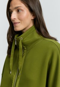 Femme portant une veste zippée olive avec un col montant et un cordon de serrage, regardant légèrement vers la gauche contre un fond clair.