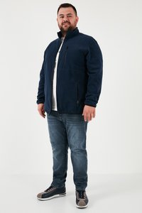 Mees seisab, kandes tumesinist zip-up jopet valge särgi peal, siniseid teksapükse ja halli-tumesiniseid tossusid, taustaks hele ühevärviline pind.