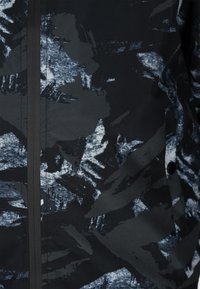 Zwart en grijs patroonstof met abstracte vormen, voorzien van een gladde textuur en een voorrits. Inclusief een knoopdetail.