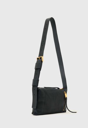 Borsa a tracolla in pelle nera con ampia tracolla regolabile, fibbia dorata, tasca frontale e cursore della cerniera dorato su sfondo grigio chiaro.