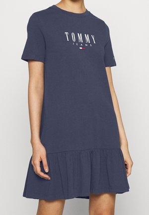 Marineblaues Kleid mit kurzen Ärmeln, Rüschensaum und "Tommy Jeans"-Logo auf der Brust, getragen von einer Person mit entspannt an den Seiten hängenden Armen.