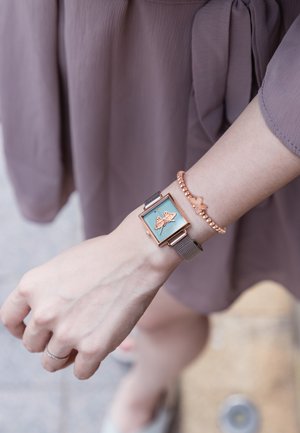 Reloj cuadrado con una esfera turquesa que presenta un diseño de mariposa, con herrajes en oro rosado y una pulsera de malla plateada, llevado junto a una pulsera de oro rosado.
