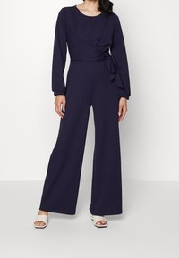 Marinblå jumpsuit med långa ärmar, samlad midja och knytdetalj. Vida ben och slätt tyg. Neutrala öppen-tå skor med klack.