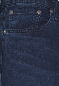 Gros plan sur une poche de jean en denim bleu foncé avec des rivets métalliques et des détails de couture visibles.