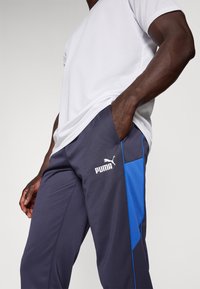 Pilki sportiniai kelnės su mėlynomis detalėmis, su Puma logotipu. Minkšta medžiaga, siaurėjantis dizainas, šoninės kišenės ir patogus prigludimas.