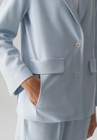 Blazer azzurro chiaro realizzato in tessuto testurizzato. Presenta rever a V, due tasche frontali e chiusura con bottone davanti. Vestibilità morbida e rilassata.
