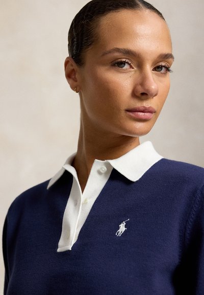 Femme aux cheveux tirés en arrière, vêtue d'un polo bleu marine avec col blanc et logo, regardant légèrement sur le côté sur un fond uni.