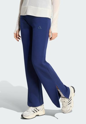 Persoon die marineblauwe uitlopende broek met zijsplitten, witte sneakers en een wit topje met lange mouwen draagt, staand op een lichte achtergrond.