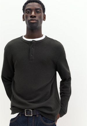 Jeune homme aux cheveux courts et bouclés portant une chemise henley noire à manches longues par-dessus un maillot blanc et un jean bleu foncé avec une ceinture noire.