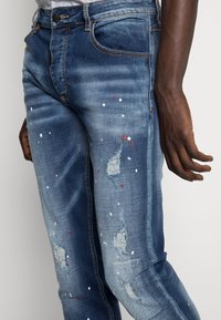 Pantalones vaqueros de mezclilla azul con un diseño desgastado, que presentan rasgaduras y salpicaduras de colores en rojo y blanco. Textura suave, corte ajustado.