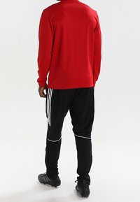 Rode sportieve top met lange mouwen, gecombineerd met zwarte broek met witte strepen en accenten, gedragen met zwarte voetbalschoenen.