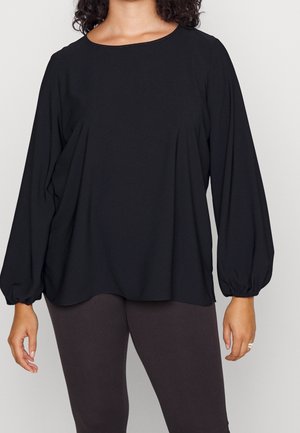 Femme portant un chemisier noir ample à manches longues avec poignets froncés et un pantalon ajusté foncé sur un fond clair uni.