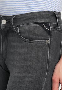 Dark gray denim jeans with a slim fit, featuring a silver button, subtle whiskering, and a black tag with "REPLAY" stitched on the waistband.
