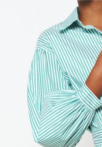 Turkusowa i biała koszula w paski z kołnierzykiem, bufiastymi rękawami i marszczonymi mankietami, wykonana z lekkiego materiału.