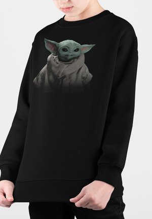 Felpa nera con una grafica che raffigura un personaggio verde con grandi orecchie, vestito con un mantello. Il materiale sembra morbido e ha una vestibilità ampia.