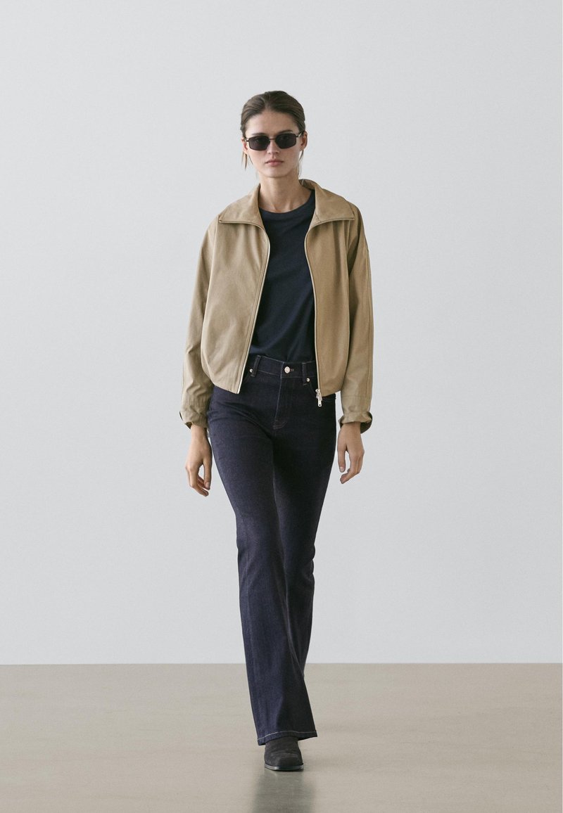 Krämfärgad kort jacka, marinblå t-shirt, mörkblå utsvängda jeans och svarta solglasögon. Jacka i slät tyg med ståkrage.