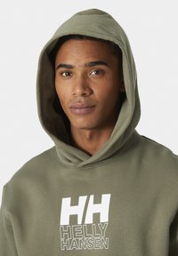 Sudadera de color verde oliva con un gran logo blanco de "HH". Presenta una capucha con cordón y tejido suave y texturizado. Primer plano de la parte superior del cuerpo.