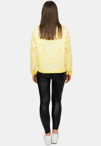 Geel gebreid sweater met witte luipaardprint, gecombineerd met zwarte glanzende legging en witte sneakers, vanuit de achterkant bekeken.