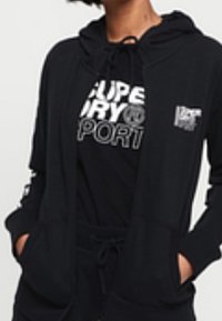 Chaqueta con capucha negra de cremallera, hecha de una tela suave, que cuenta con un bolsillo frontal y capucha con cordón, llevada sobre una camiseta gráfica negra.