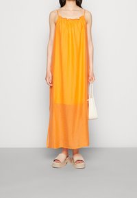 Robe maxi orange avec un col froncé et des fines bretelles ; le mannequin tient un petit sac blanc et porte des sandales à plateforme beiges.