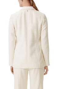 White textured blazer with a fitted design and notched collar. Features a subtle back branding detail. Long sleeves and tailored silhouette.