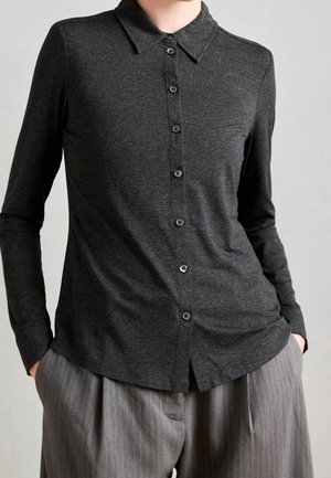 Ciemnoszara, koszula z długim rękawem na guziki z kołnierzykiem, noszona wpuszczona do szarych spodni w cienkie prążki, ręce w kieszeniach, neutralne tło.