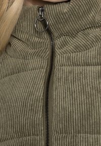 Chaqueta de pana verde oliva con un alto cuello, textura acanalada y un cierre central de cremallera negra con un tirador circular.