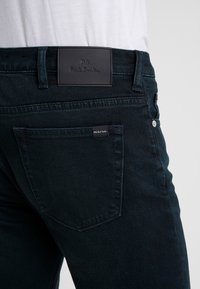 Dark wash denim jeans with a slim fit, featuring a black leather brand patch at the waistband and a small logo tag on the back pocket.