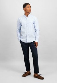 Camicia a maniche lunghe azzurro chiaro con colletto alla coreana, bottoni frontali e taschino sul petto, abbinata a pantaloni scuri e scarpe marroni.