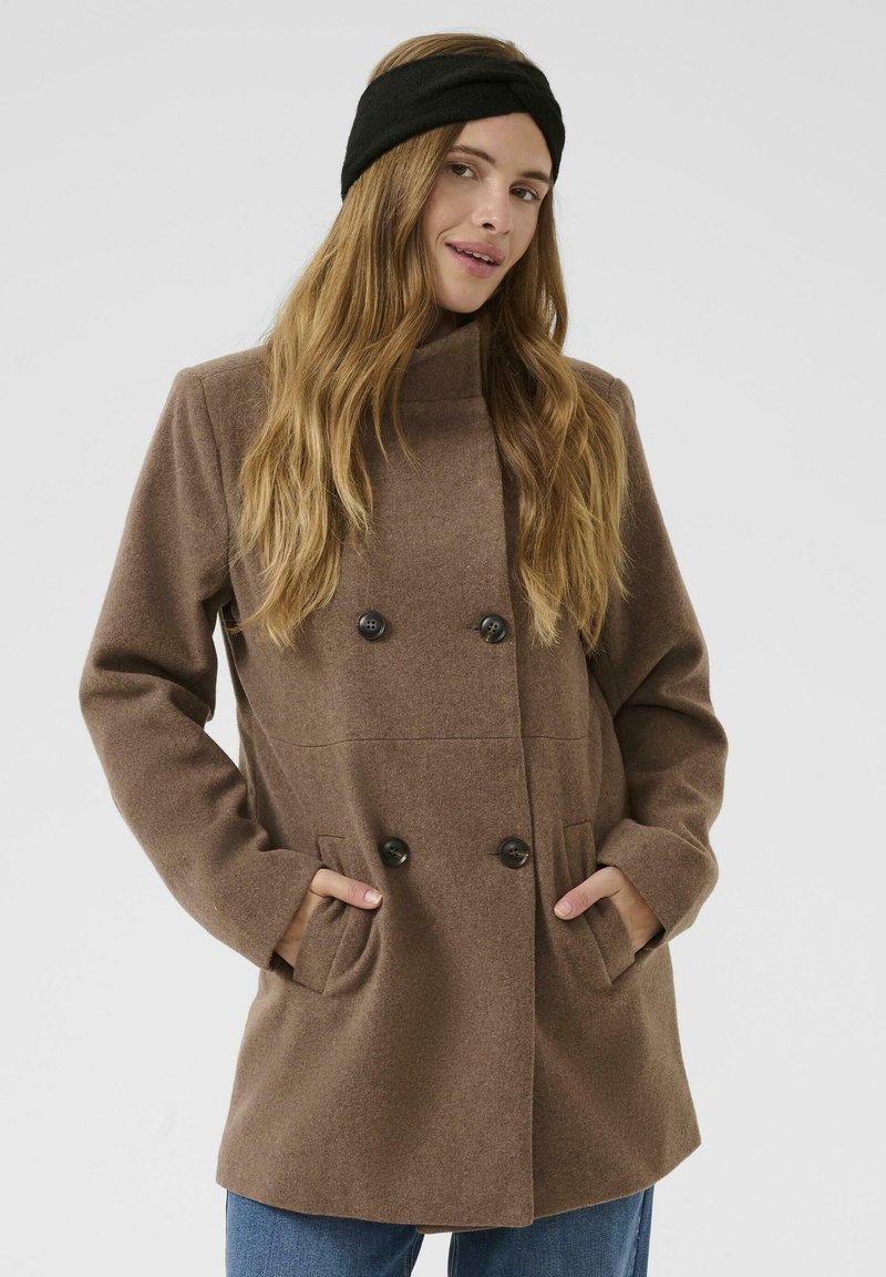 Brun dobbeltradet frakke lavet af uldblanding, med høj krave, sidelommer og store sorte knapper. Model iført en sort pandebånd.