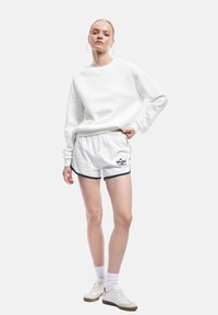 Sudadera blanca con mangas raglán, combinada con pantalones cortos blancos que tienen un acento en azul marino y texto. El modelo lleva zapatillas blancas y calcetines.