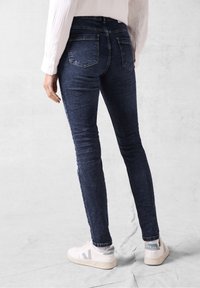 Jeans skinny bleu marine avec une texture en denim lisse, dotés d'une taille haute et de deux poches arrière. Portés avec des sneakers blanches.
