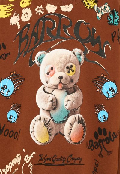 Tissu marron avec un ours en peluche cousu ayant des boutons d'yeux dépareillés, des patchs colorés, et des graphismes et textes de style bande dessinée autour.