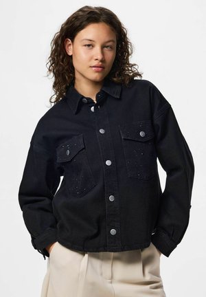 Tânără cu păr creț, purtând o geacă neagră cu nasturi și buzunare la piept și pantaloni de culoare deschisă, pozând cu mâinile în buzunare.