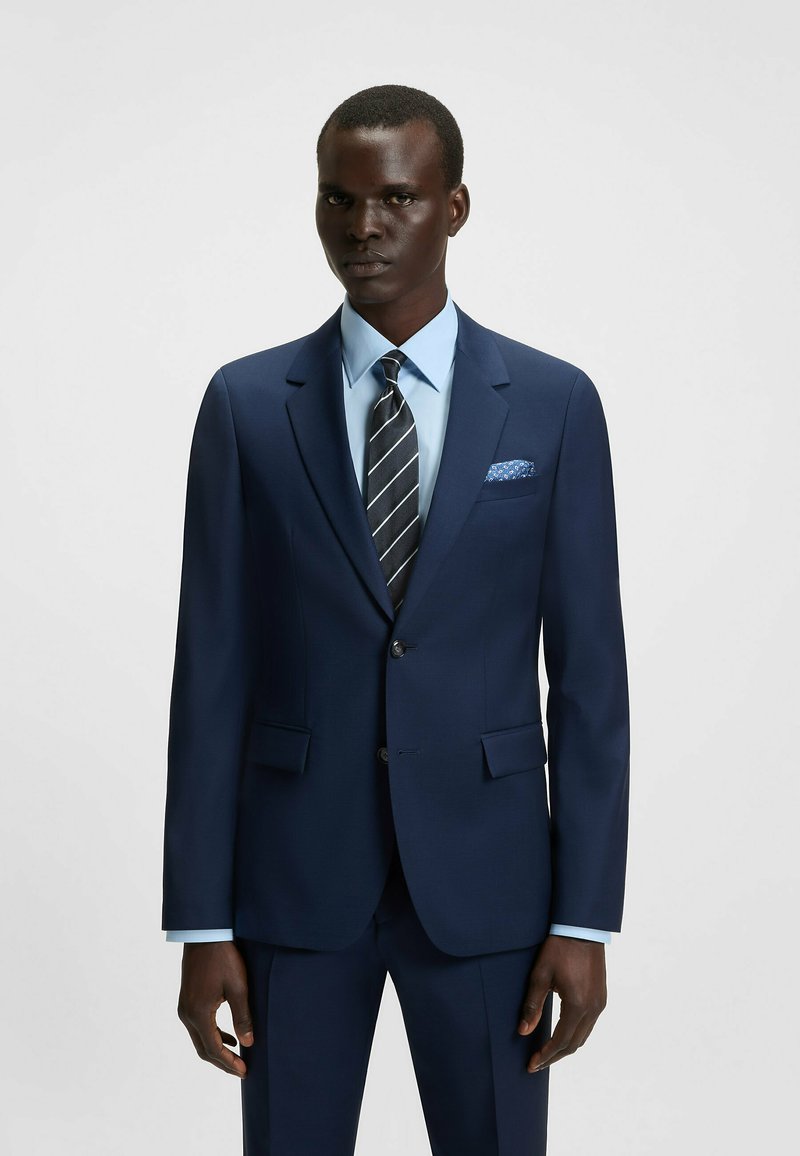 Tummansininen puku, jossa on lovelliset kaulukset, kaksi nappia ja pehmeä rakenne. Yhdistetty vaaleansiniseen paitaan ja musta-valkoiseen raidoitettuun solmioon.