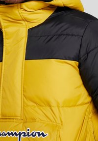 Gul och svart vadderad jacka med en framficka, tydliga sömmar och huva. Inkluderar en vit "Champion"-logga på framsidan.