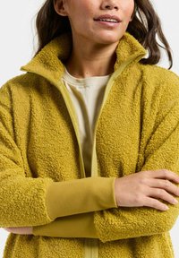 Veste en polaire jaune avec une surface texturée et douillette, col montant et poignets extensibles. En dessous, un haut clair à col rond.