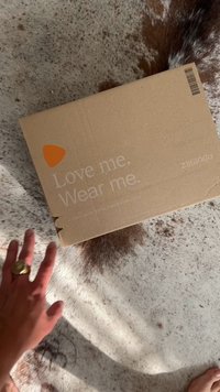 Καφέ χαρτονένιο κουτί με εκτυπωμένο κείμενο "Αγάπησέ με. Φόρεσέ με." και ένα πορτοκαλί λογότυπο Zalando. Τοποθετημένο σε επιφάνεια με ανοιχτόχρωμη υφή.